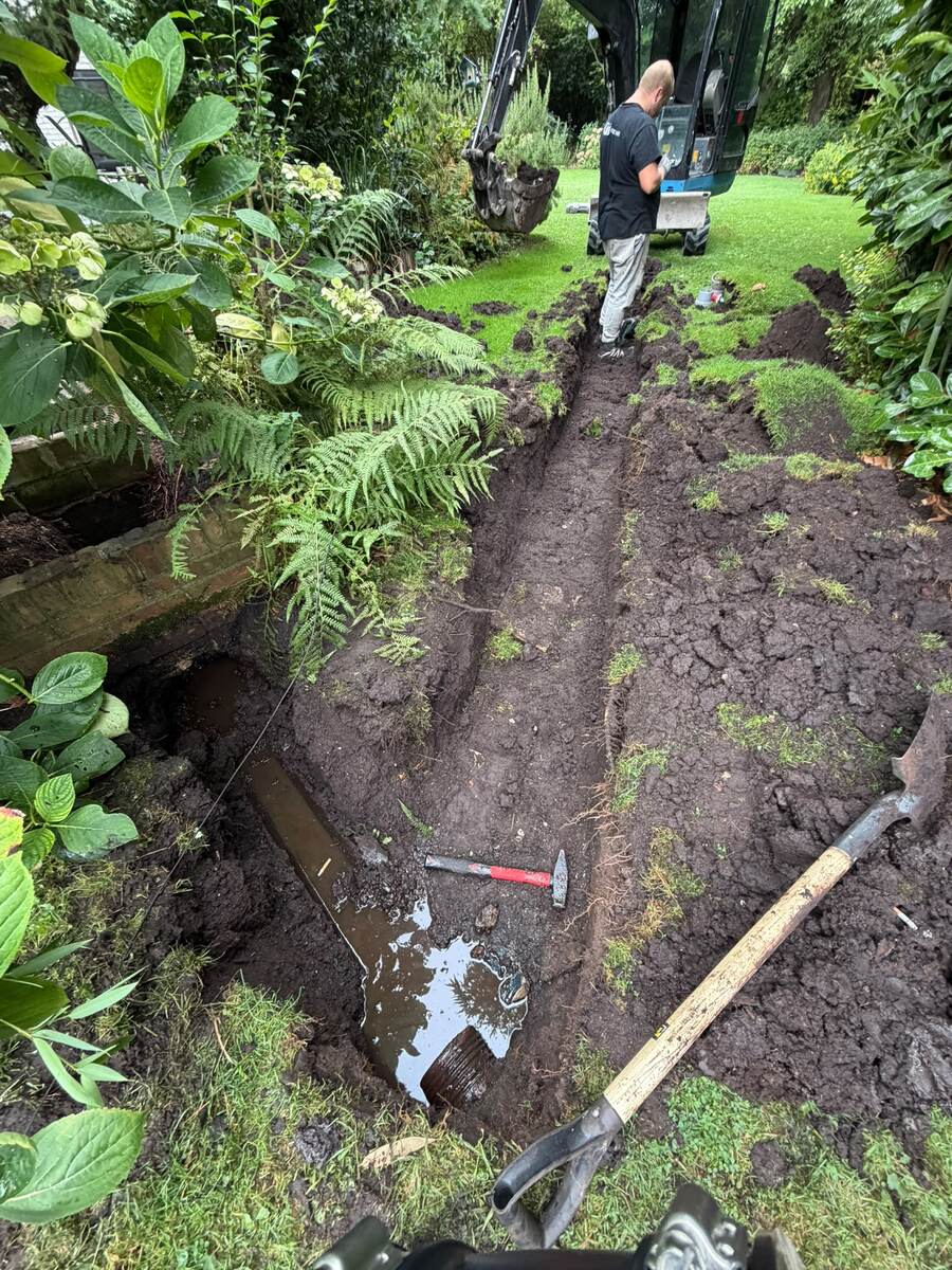 Minigraver naast een uitgegraven sleuf in de tuin waarin de oude rioolbuis blootligt